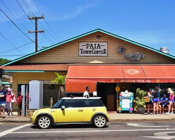 Yellow car stopped outside Paia Town Center as tourists stroll along the sidewalk.