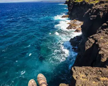 Person's feet hanging over the edge of a cliff overlooking the ocean.