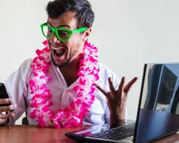 Man wearing lei and green glasses excited by his phone. He's sitting at a desk and there's a laptop open.