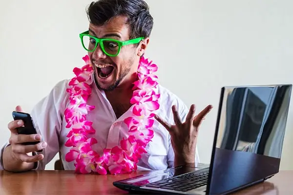 Man wearing lei and green glasses excited by his phone. He's sitting at a desk and there's a laptop open. 