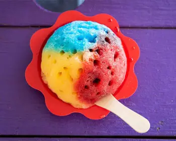 Red, yellow, and blue shave ice on purple wood table.