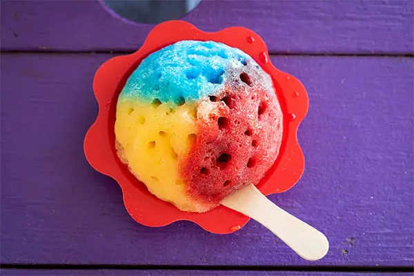 Red, yellow, and blue shave ice on purple wood table.  