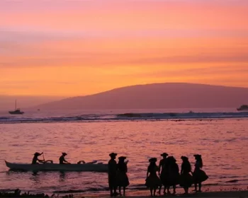 People in traditional Hawaiian garb standing on the beach at sunset.