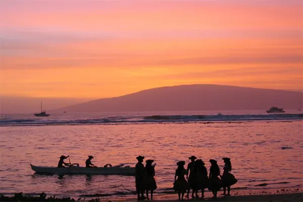 People in traditional Hawaiian garb standing on the beach at sunset. 