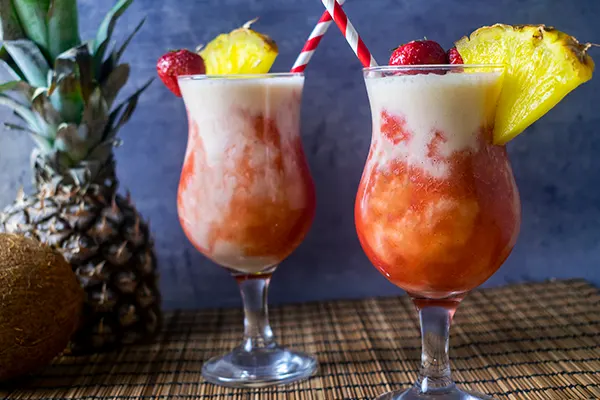 Two lava flow drinks garnish with pineapple on a table cloth and a pineapple behind them. 