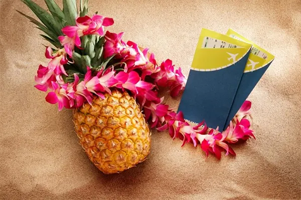 Pineapple draped in a lei next to two plane tickets on the beach. 