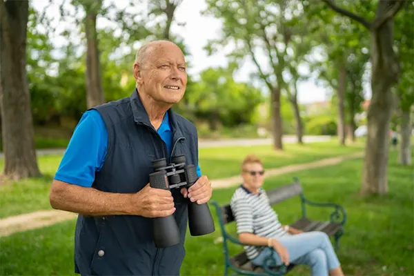 A senior man in the park holding binoculars by his chest.