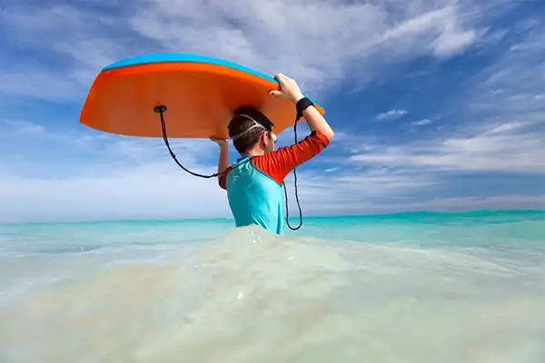Boy holding boogie board over his head as he wades into the ocean. 